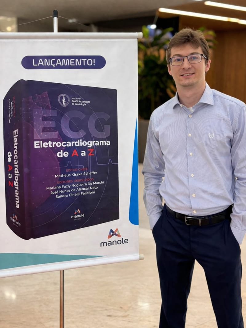Dr. Matheus Kiszka Scheffer ao lado do banner de lançamento do livro Eletrocardiograma de A a Z